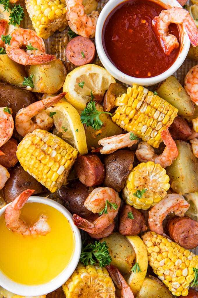 A sheet pan with shrimp, corn, sausage and potatoes. Two bowls on either side with cocktail sauce and butter.