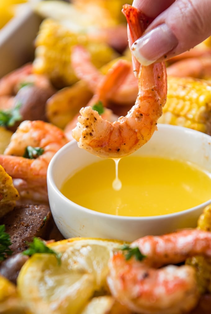 A shrimp being dipped into a bowl of butter, surrounded by sausage and corn on the cob.