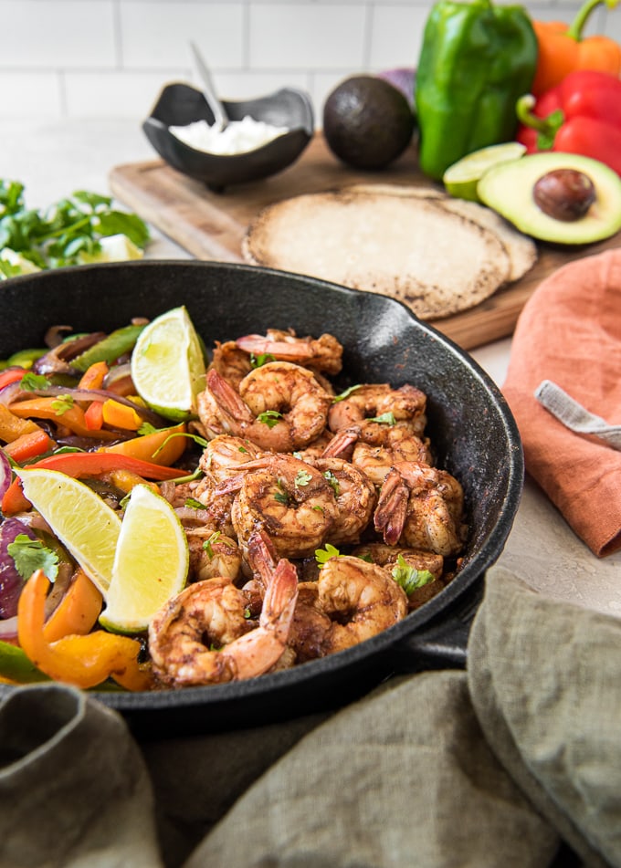 A skillet with shrimp, peppers, onions and lime surrounded by tortillas,m avocado and napkins