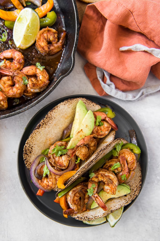 A couple of shrimp fajitas on a plate and a cast iron skillet with shrimp, peppers and lime.