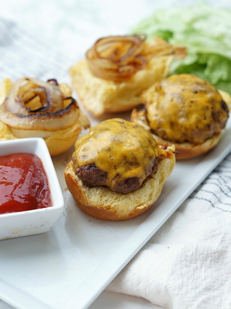 Grilled Hamburger Sliders sitting on a white platter next to a dish of ketchup. 2 of the sliders are topped with grilled onions and all are topped with yellow cheese.