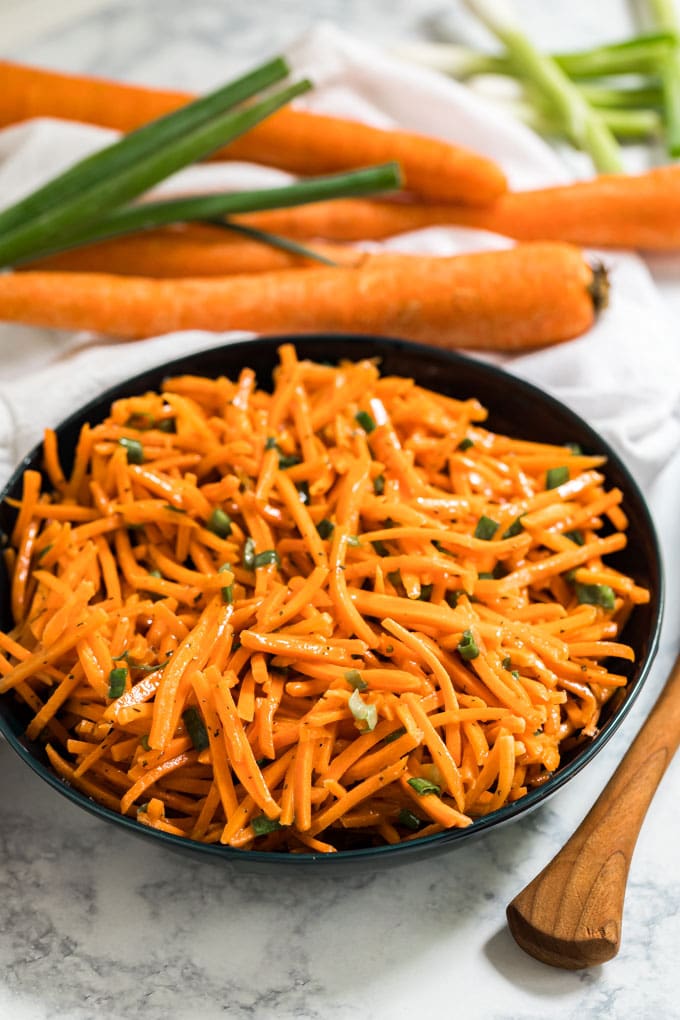A bowl of shredded carrot salad with carrots behind it.