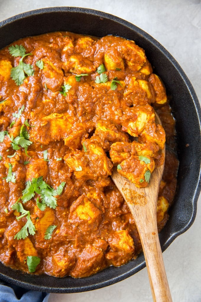 A large cast iron skillet filled with chicken and curry sauce. A wooden spoon sits in the pan.