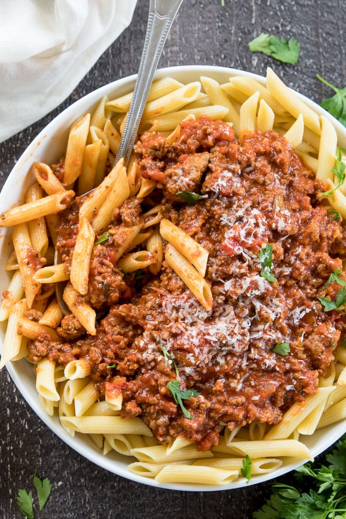 A large white bowl with penne pasta and bolognese meat sauce with a spoon to stir it up.