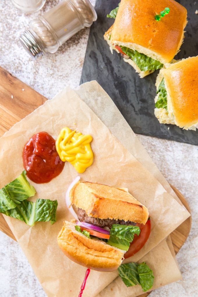 2 hamburger sliders, one turned on it's side, resting on parchment paper and sitting next to dollops of ketchup and mustard. 2 sliders are sitting on a black cutting board and a bottle of pepper is resting on it's side.