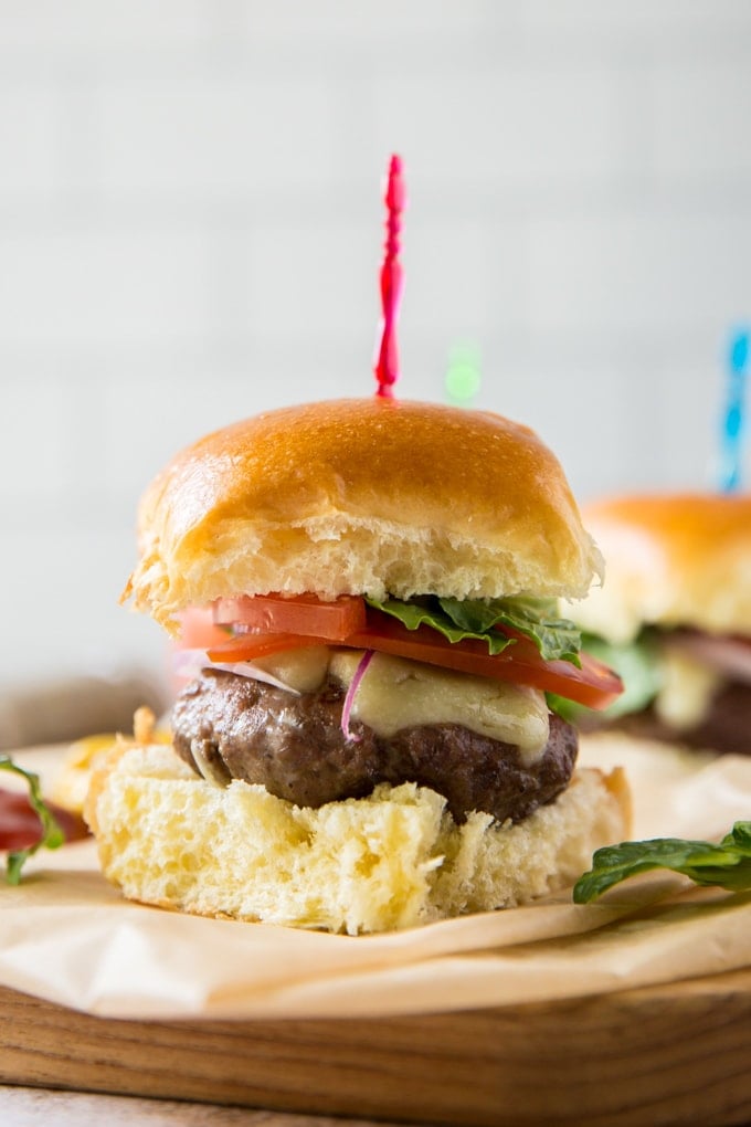 a close up of a hamburger slider topped with cheese, tomato and lettuce with a red toothpick sticking out of it. sitting on parchment paper on top of a wood board.