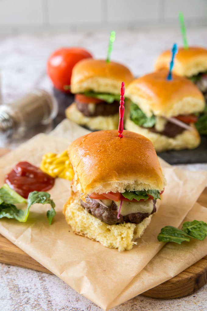 4 mini slider hamburgers with colored toothpicks sitting on brown parchment paper on top of a wood cutting board.