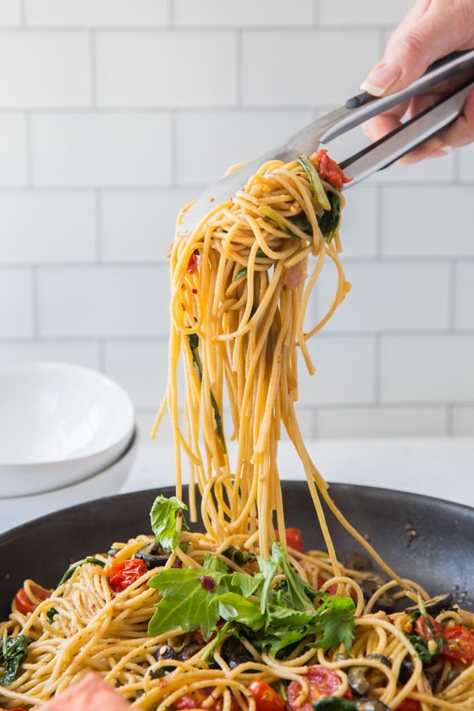 Pasta Puttanesca is a light meal of fresh pasta with lemon juice, tomatoes, spinach and arugula and a robust saltiness from capers and olives. pasta picked up with tongs from a large skillet