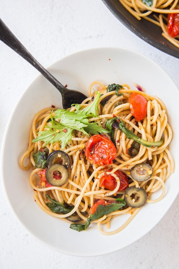 Pasta Puttanesca is a light meal of fresh pasta with lemon juice, tomatoes, spinach and arugula and a robust saltiness from capers and olives. A white bowl full of pasta puttanesca