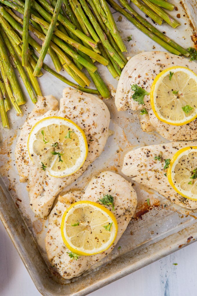 An overhead shot of chicken breasts with lemon slices on top next to asparagus on a sheet pan