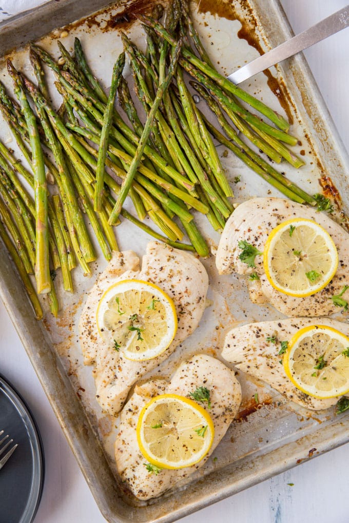 An overhead shot of chicken breasts with lemon slices and asparagus on a sheet pan.