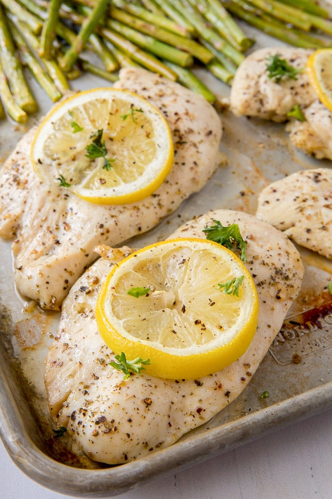 A close up of chicken breasts with lemon slices and herbs on a sheet pan with asparagus in the background