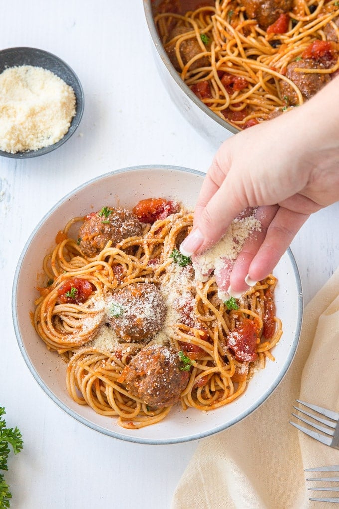 a hand sprinkling parmesan cheese over a bowl of spaghetti and meatballs, a dish of parmesan, a pan of spaghetti, a fork, parsley