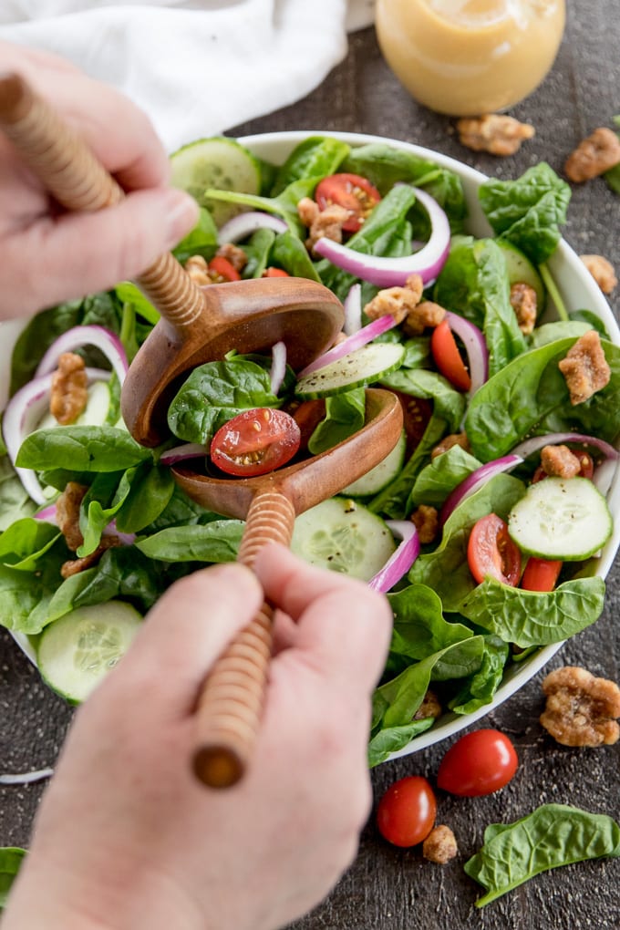 Hands reaching in with salad tongs to toss spinach with cucumber and tomatoes