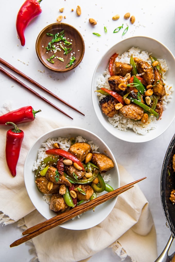 An overhead image of szechuan chicken and rice in two bowls with chopsticks and chile peppers.