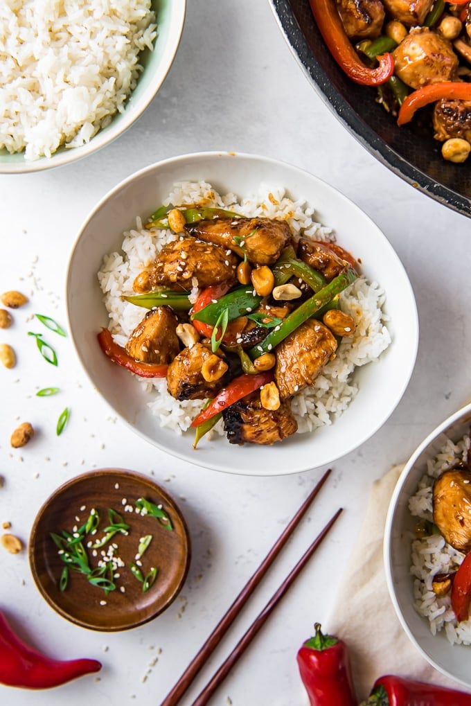 A small white bowl with white rice and szechuan chicken with peanuts, surrounded by other bowls of the same, and chopsticks.