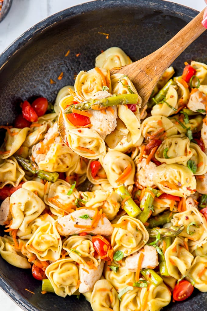 A large skillet with a wooden spoon and tortellini, chicken and vegetables