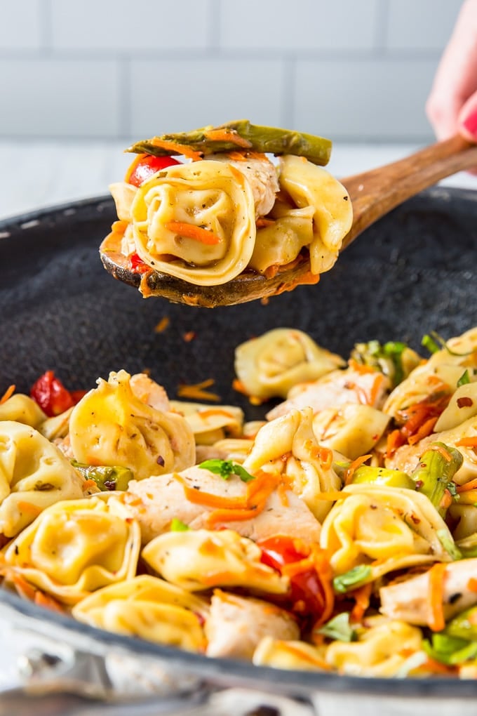 A wooden spoon holding a helping of tortellini salad over a large skillet of the same.