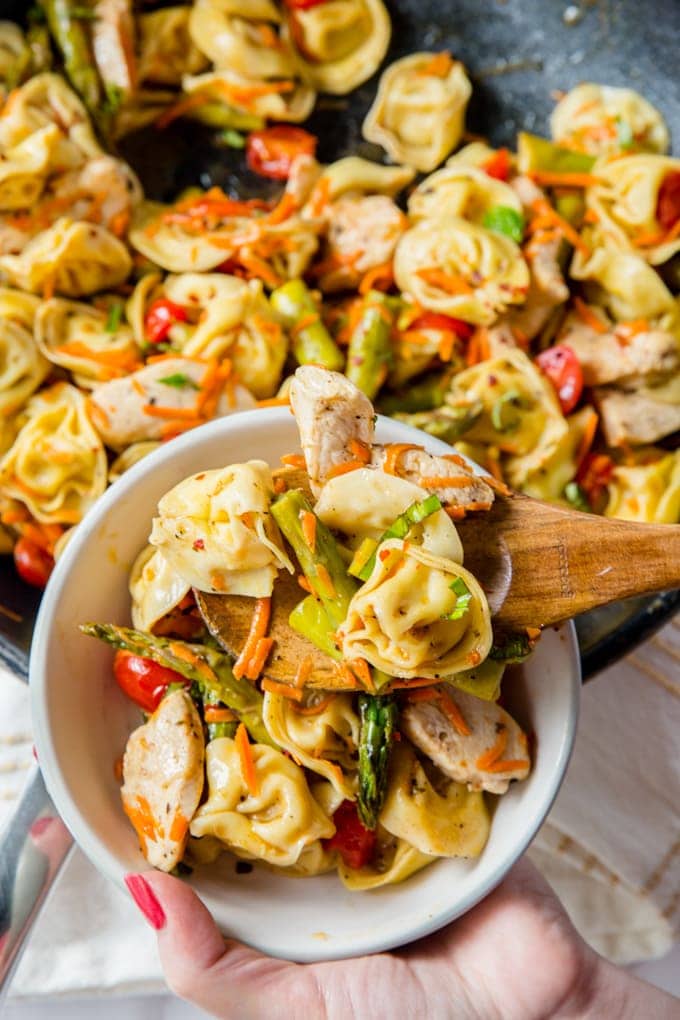 A hand holding a white bowl while a spoon fillis the bowl with tortellini salad from a skillet.