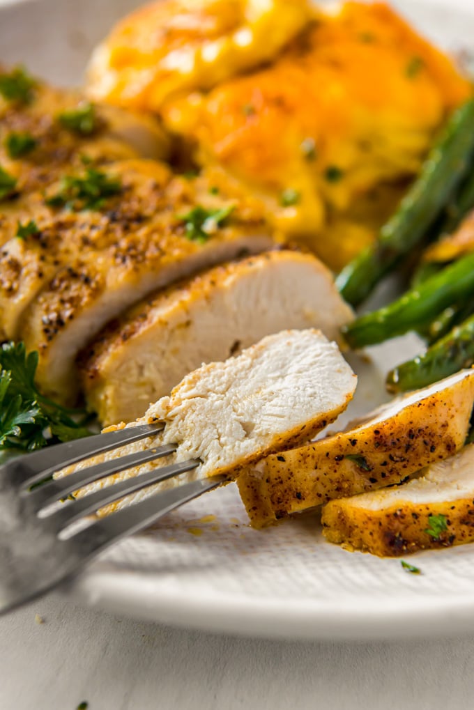 A piece of chicken breast sliced on a plate, cheesy potatoes, green beans, a fork with a piece of chicken