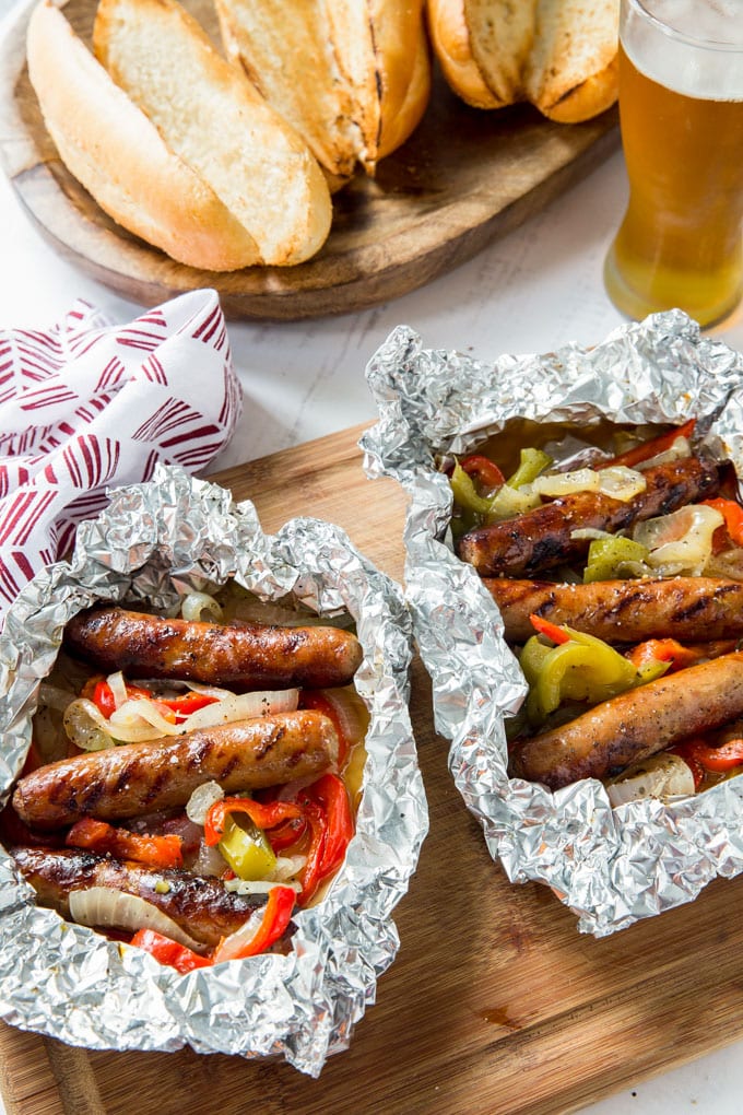 An overhead image of sausage and peppers in foil, toasted rolls and a glass of beer on wood cutting boards