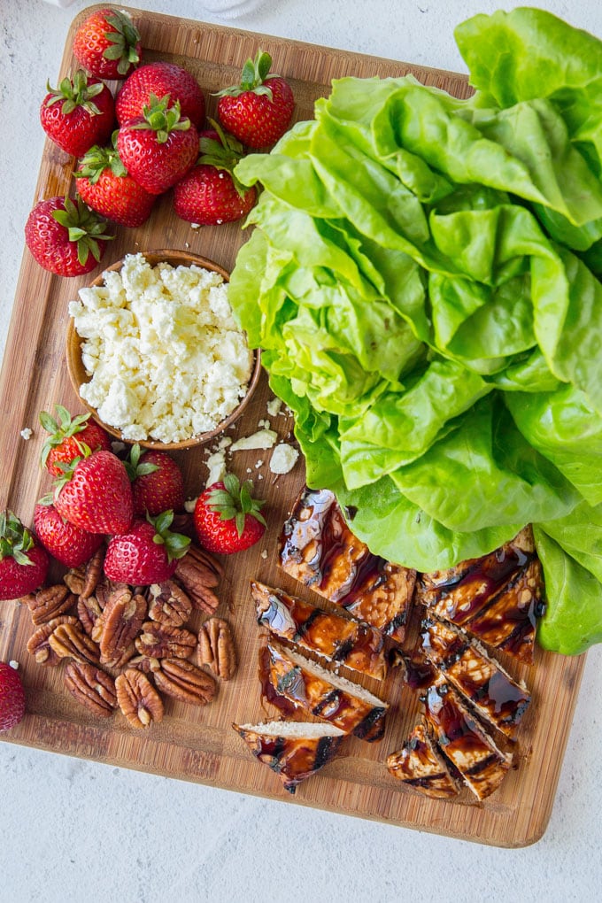Grilled Balsamic Chicken in a tangy orange balsamic marinade with strawberries, pecans and Feta. These lettuce wraps are a light and filling summer meal. Lettuce leaves, strawberries, cheese and balsamic chicken spread on a cutting board - ingredients to make balsamic chicken lettuce wraps