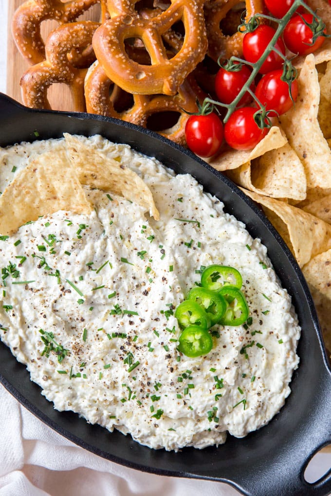 An overhead shot of jalapeno artichoke dip in a black serving dish and surrounded by pretzels, tomatoes and tortilla chips