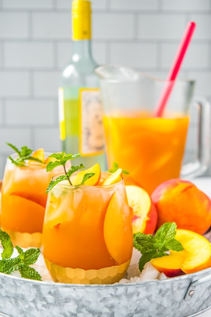 Side view of a tray of glasses filled with peach sangria, a bottle of white wine and a pitcher of sangria