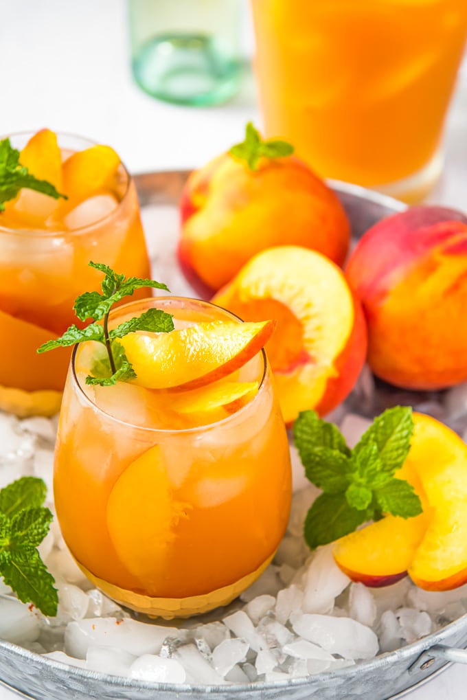 An overhead image of a glass of peach sangria, plus a pitcher and some fresh peaches and crushed ice.