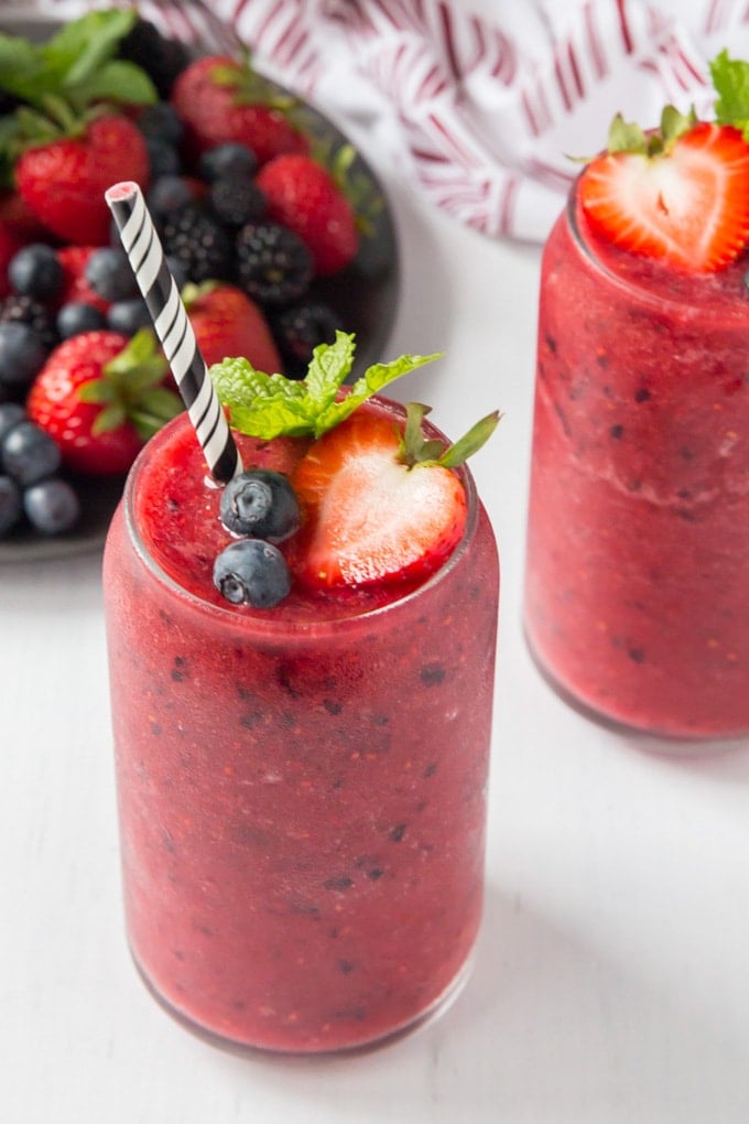 An overhead shot of two glass of berry smoothies topped with a black and white straw and fresh berries; there is a plate of fresh strawberries and blueberries in the background.