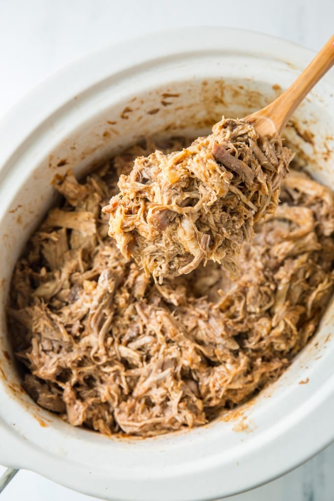 A slow cooker filled with shredded pork and a wooden spoon