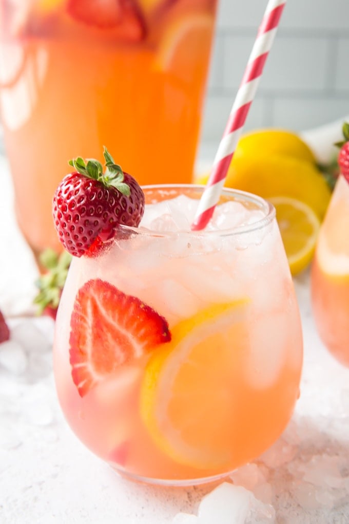 A close up of a glass of strawberry lemonade with a red and white straw and fruit garnishes