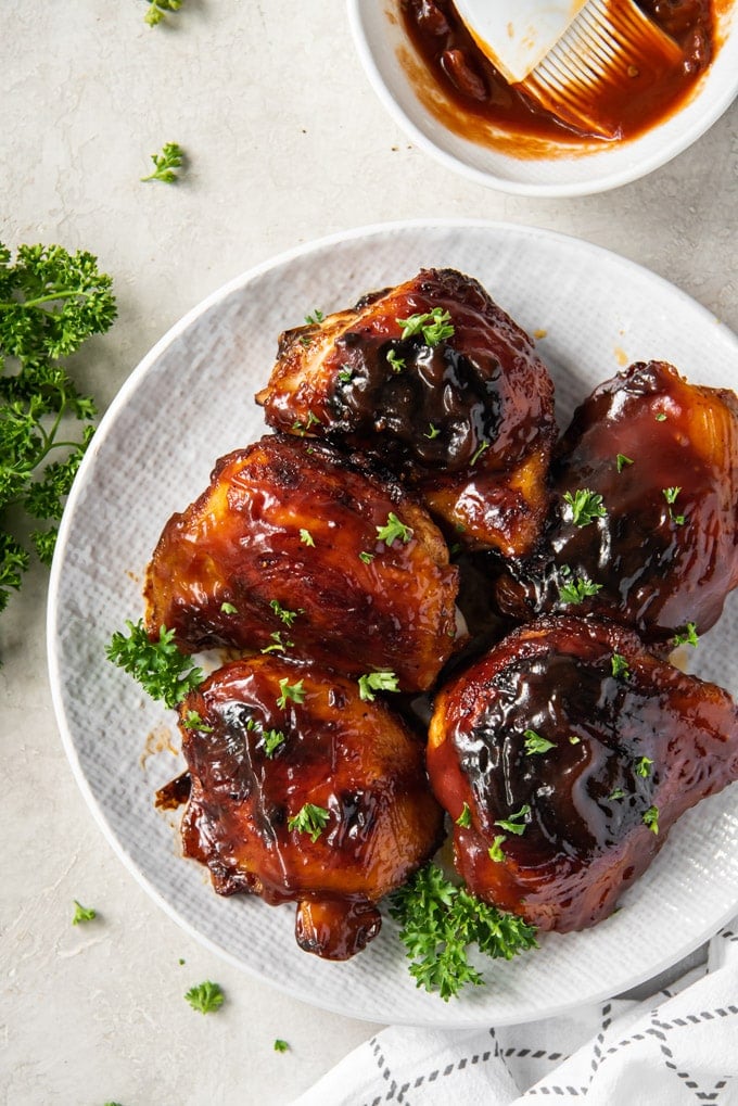 An overhead image of bbq baked chicken thighs on a white plate net to a bowl of bbq sauce