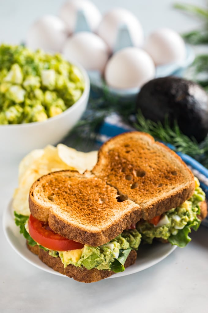 An avocado egg salad sandwich on a plate with chips.