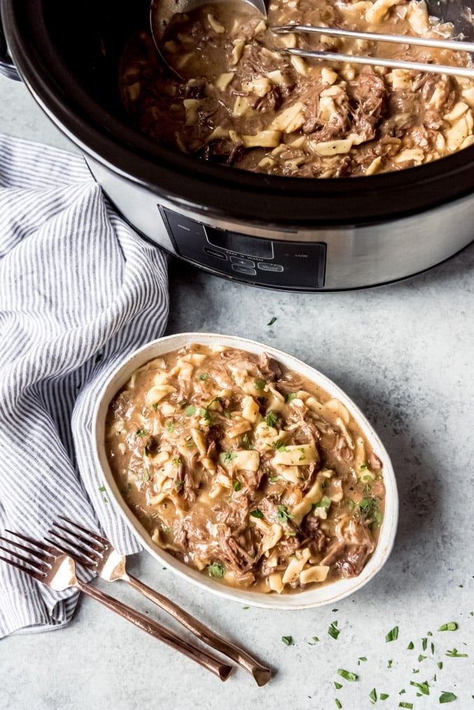 An overhead shot of a crock pot or slow cooker with beef and noodles inside, next to a dish filled with the same.