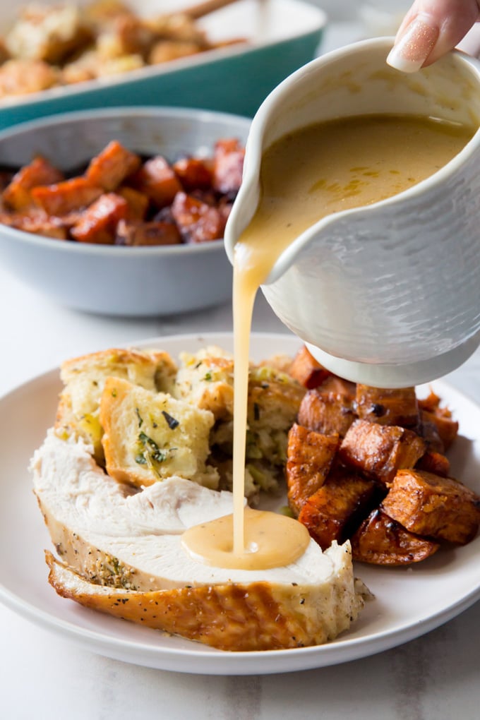 Gravy being poured from a gravy boat onto a slice of turkey breast on a plate with stuffing and sweet potatoes