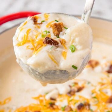 a silver ladle with potato soup, bacon, cheese and parsley, a red pot of potato soup below