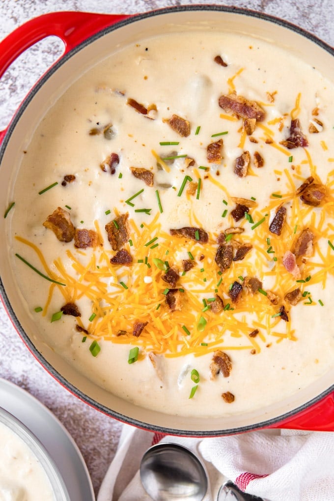 An overhead shot of a bit red pot of loaded baked potato soup topped with cheese and bacon.