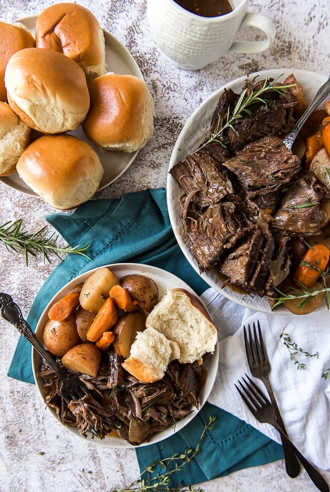 An overhead image of a plate 3 plates containing pot roast, veggies and rolls.