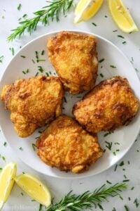 Four fried chicken thighs on a white plate, surrounded by fresh rosemary and lemon wedges.