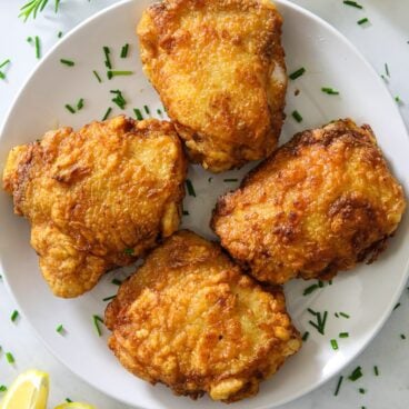 Four fried chicken thighs on a white plate, surrounded by fresh rosemary and lemon wedges.