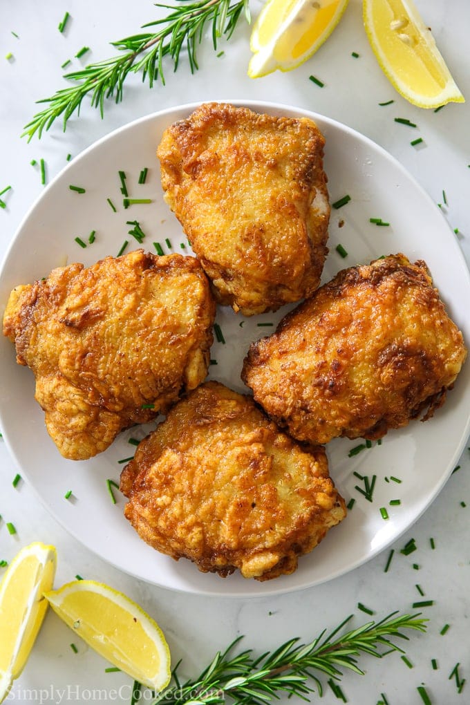 an overhead image of the best and juiciest fried chicken on a white plate with lemon wedges and chopped chives beside it