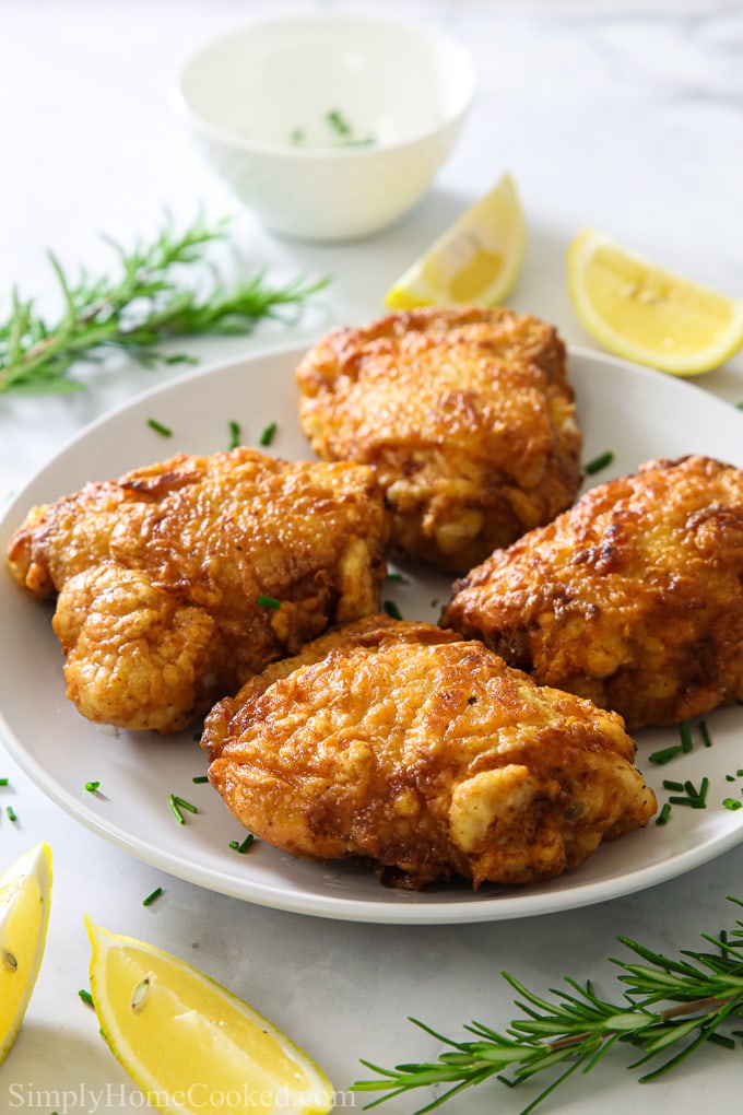 A close up image of crispy fried chicken on a white plate with fresh rosemary behind it