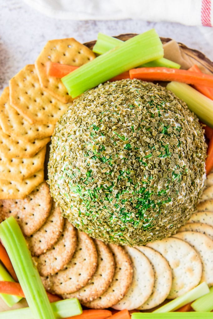 a cheese ball surrounded by crackers and veggie sticks and covered with minced chives.