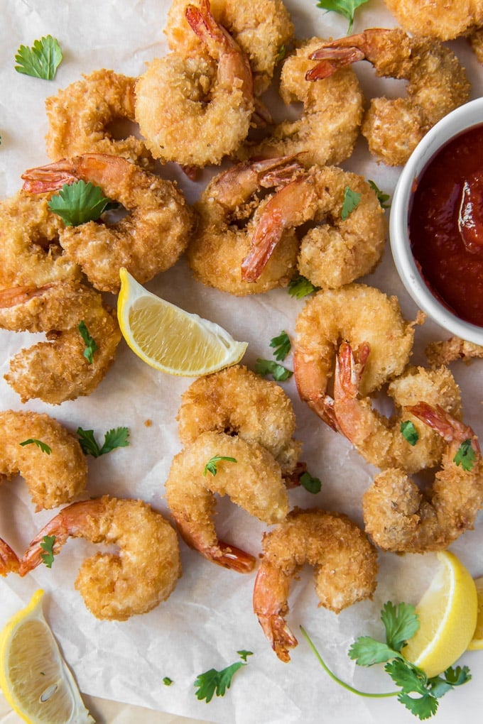 An overhead shot of a batch of fried shrimp with cocktail sauce and lemon wedges