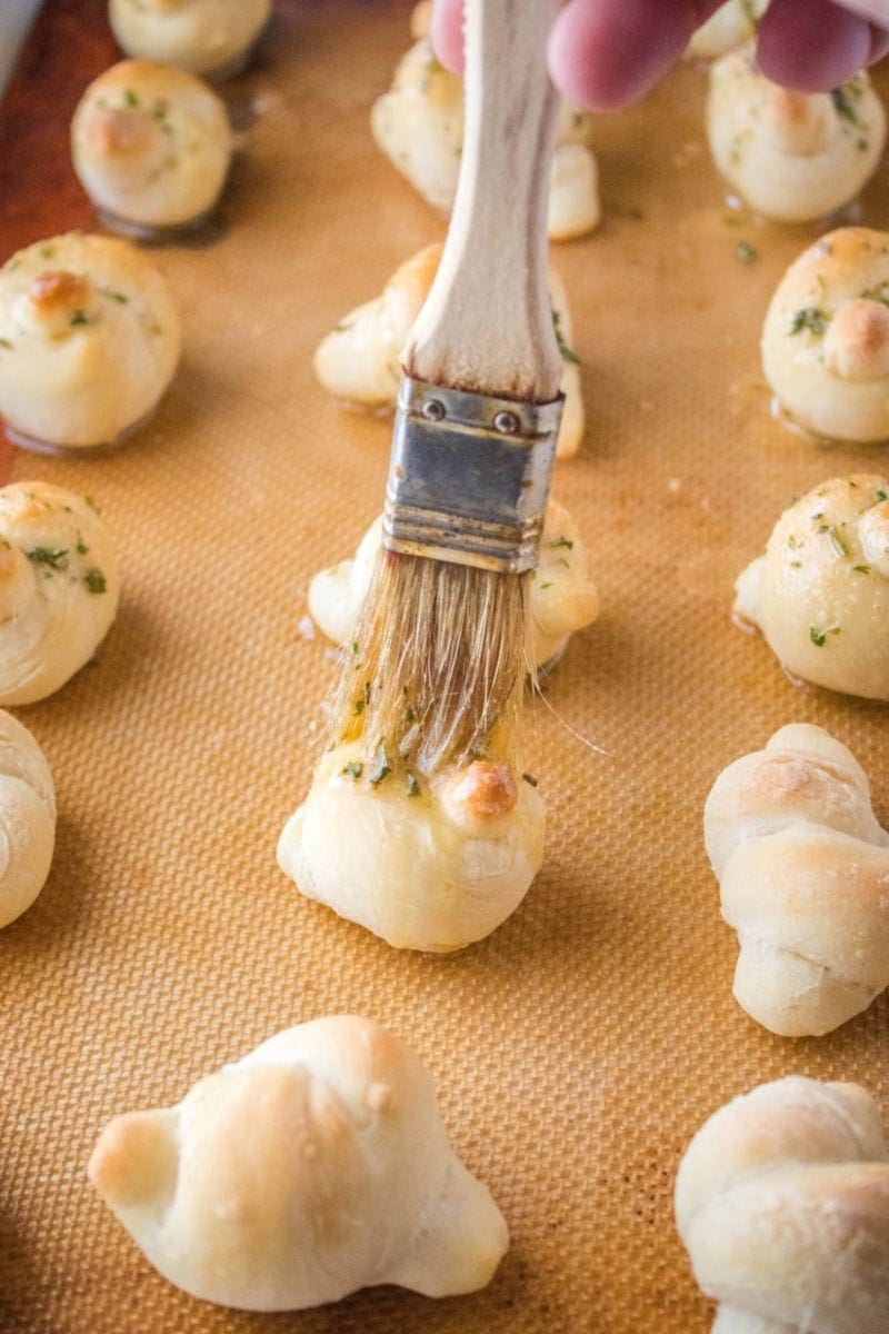 Mini Parmesan Garlic Knots are a poppable appetizer that flies off the plate! Serve them small, or make bigger knots to serve up with your favorite dinners! pastry brush coating dough knots in garlic butter