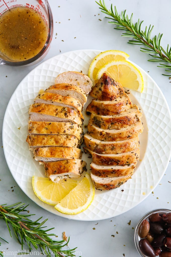 The BEST Greek Chicken Salad with with juicy grilled chicken, crunchy cucumbers, cherry tomatoes, and crumbled feta cheese. #greekchickensalad #greeksalad #chickensalald #greeksaladdressing #grilledchicken an overhead image of sliced juicy grilled chicken on a white plate with lemons and rosemary around it