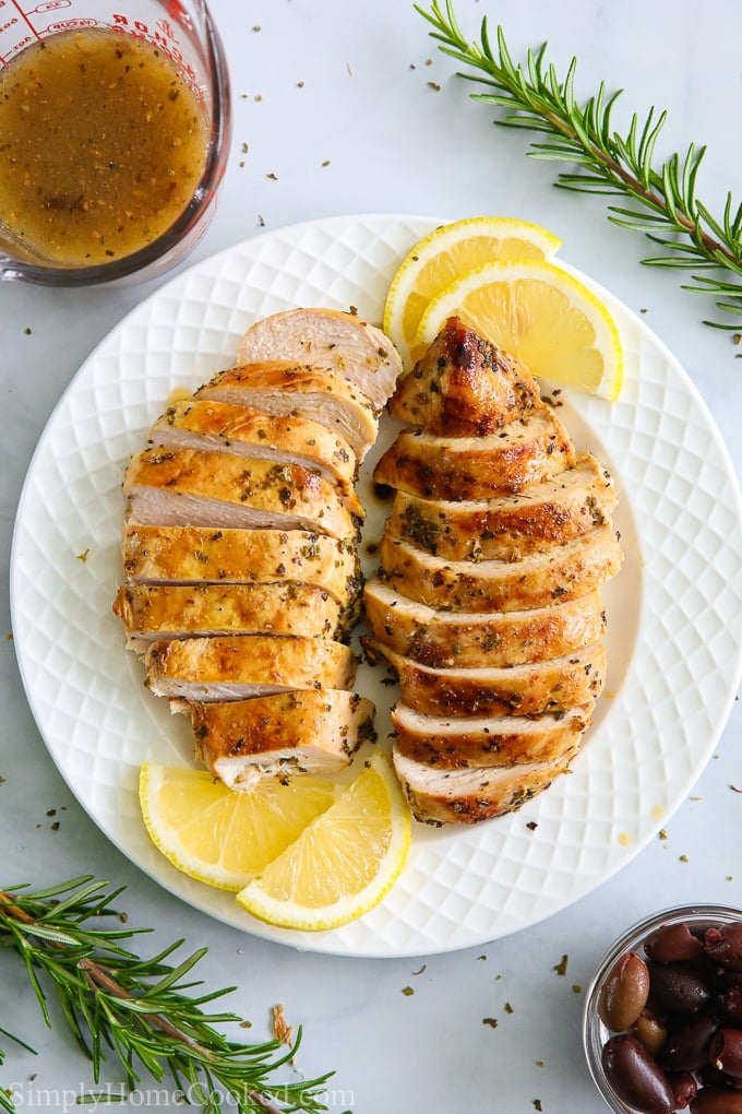 an overhead image of sliced juicy grilled chicken on a white plate with lemons and rosemary around it 