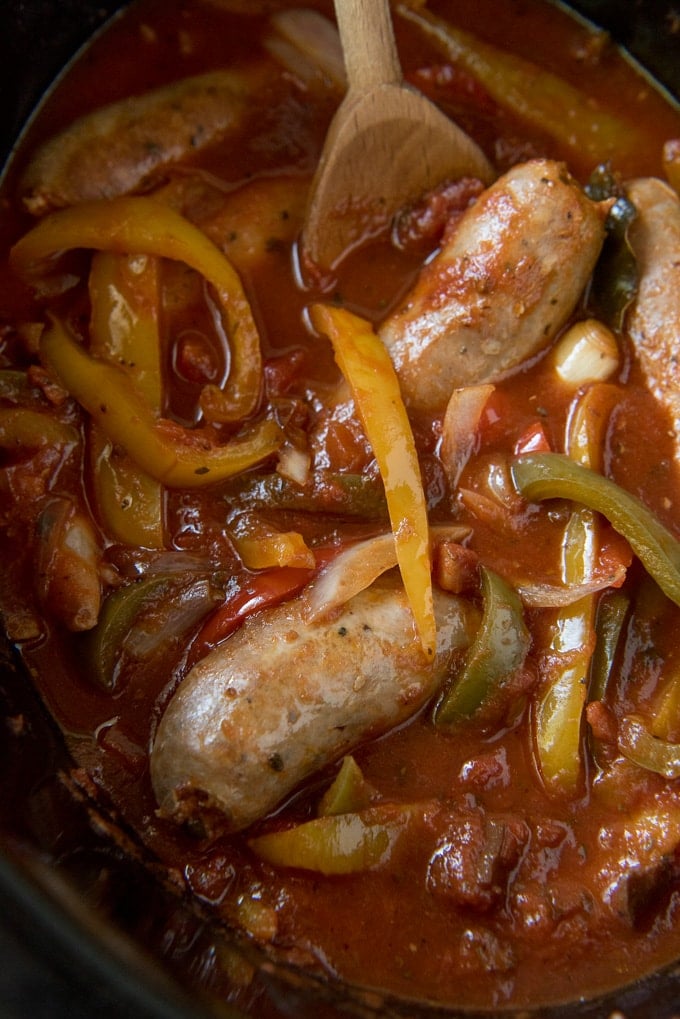 Italian sausage, peppers and onions in marinara sauce in the slow cooker with a wooden spoon.