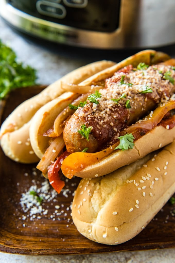 A stack of hoagie rolls with italian sausage adn peppers with a slow cooker in the background.